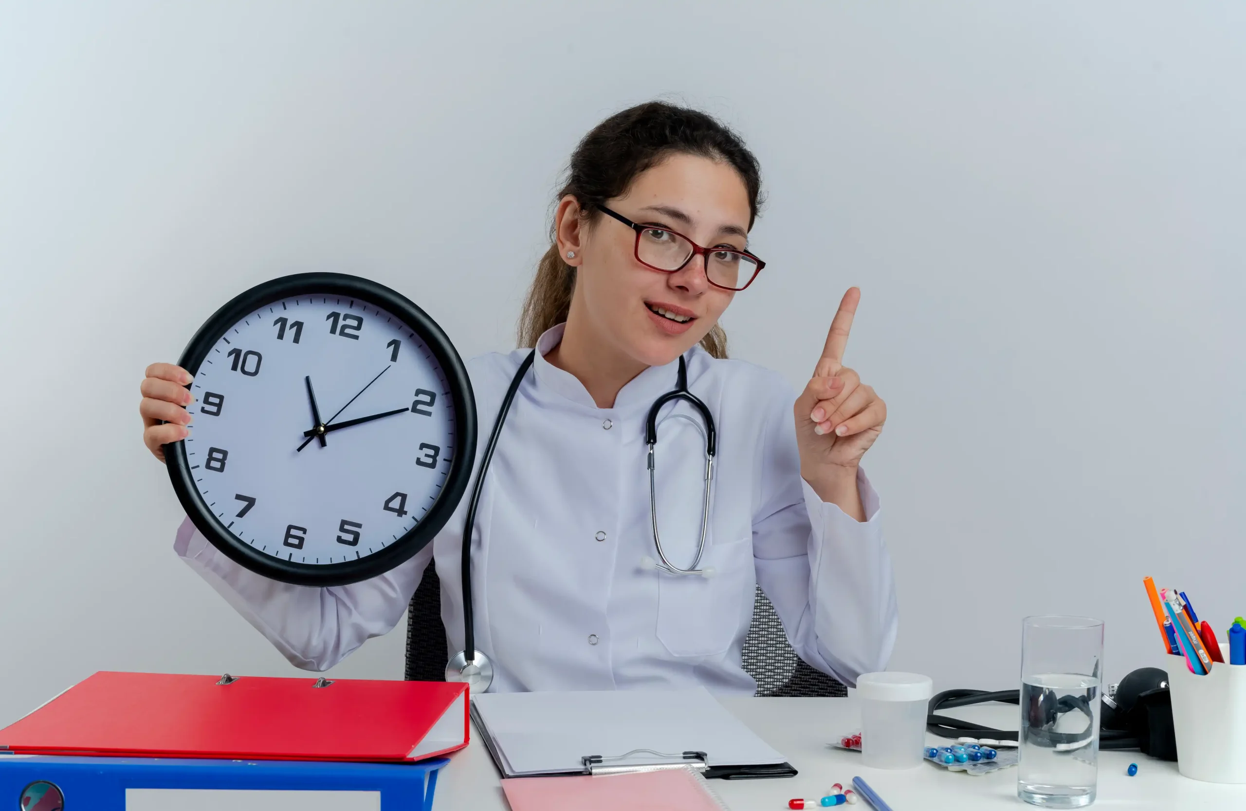 impressed young female doctor wearing medical robe stethoscope glasses sitting desk with medical tools looking holding clock raising finger isolated